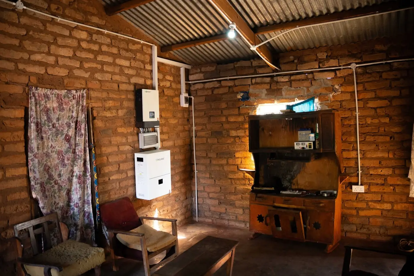 A wall-mounted inverter and battery inside a Lazaro Village home, beside a lit bulb